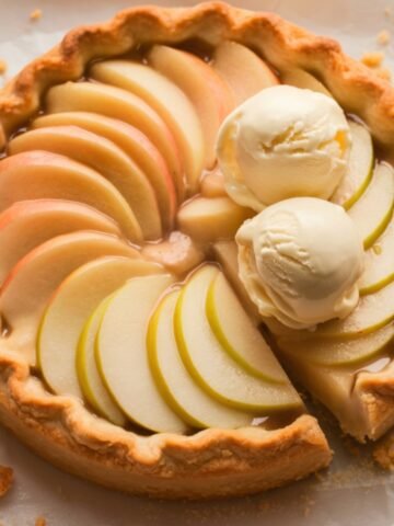 A golden Puff Pastry Apple Tart with a flaky, buttery crust, filled with beautifully arranged thin apple slices. The tart is topped with a glossy glaze and served with two scoops of vanilla ice cream, slightly melting over the warm apple filling. Crumbly edges and a spoon nearby hint at a delicious homemade dessert, perfect for any occasion.